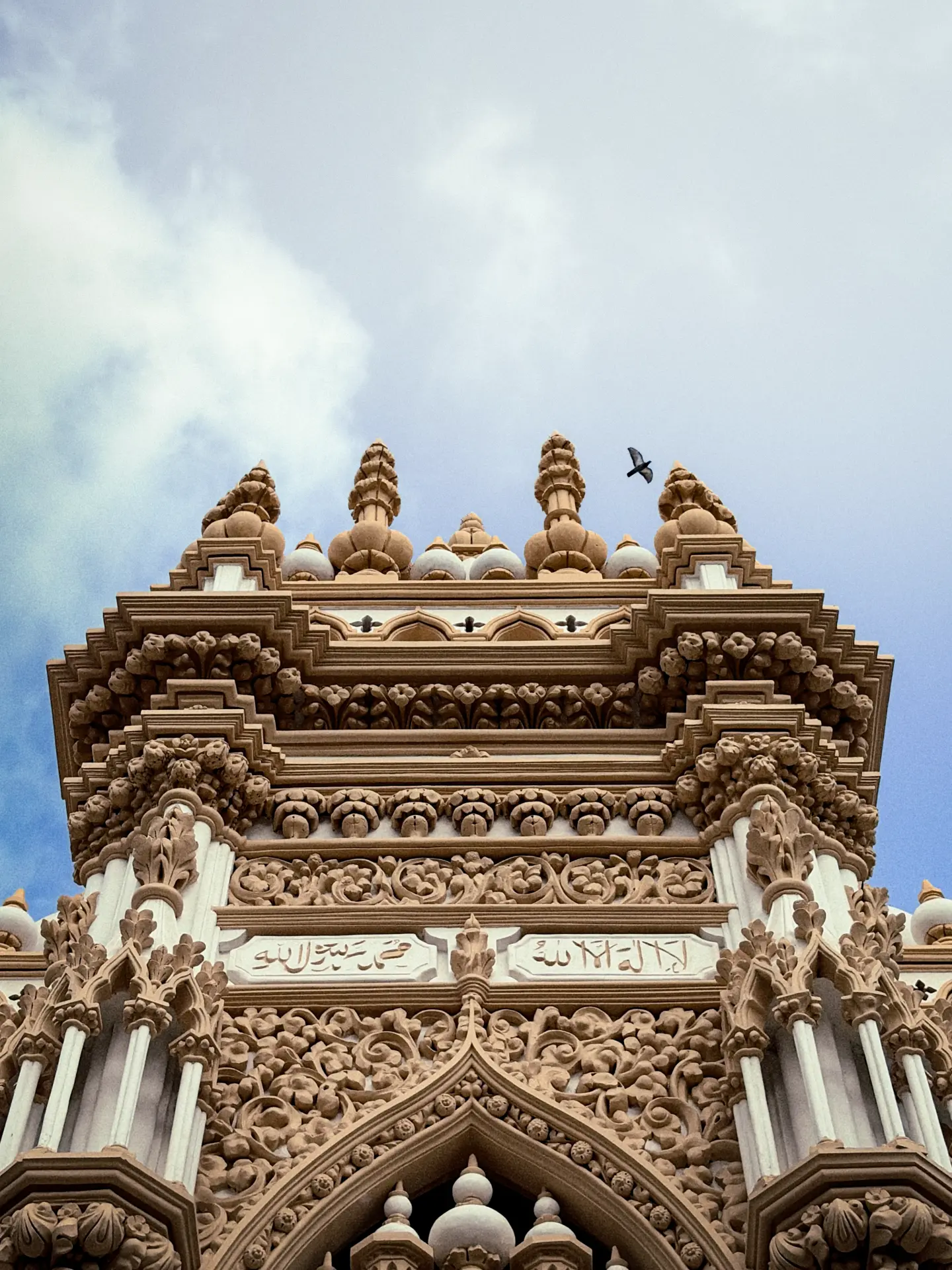 Upward view of an intricately carved sandstone Islamic tower with floral relief work, white marble inlay and Arabic calligraphy panels, a bird flying past the blue and cloud-streaked sky above.