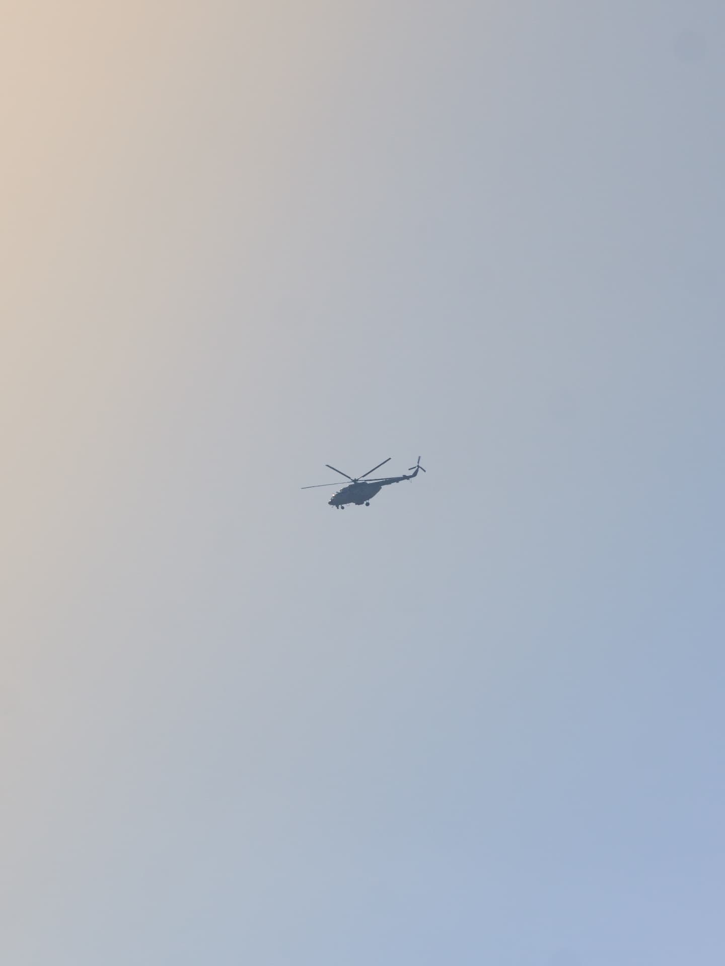 A distant silhouetted military transport helicopter flying across a vast hazy blue and warm-toned evening sky with no other elements in frame.