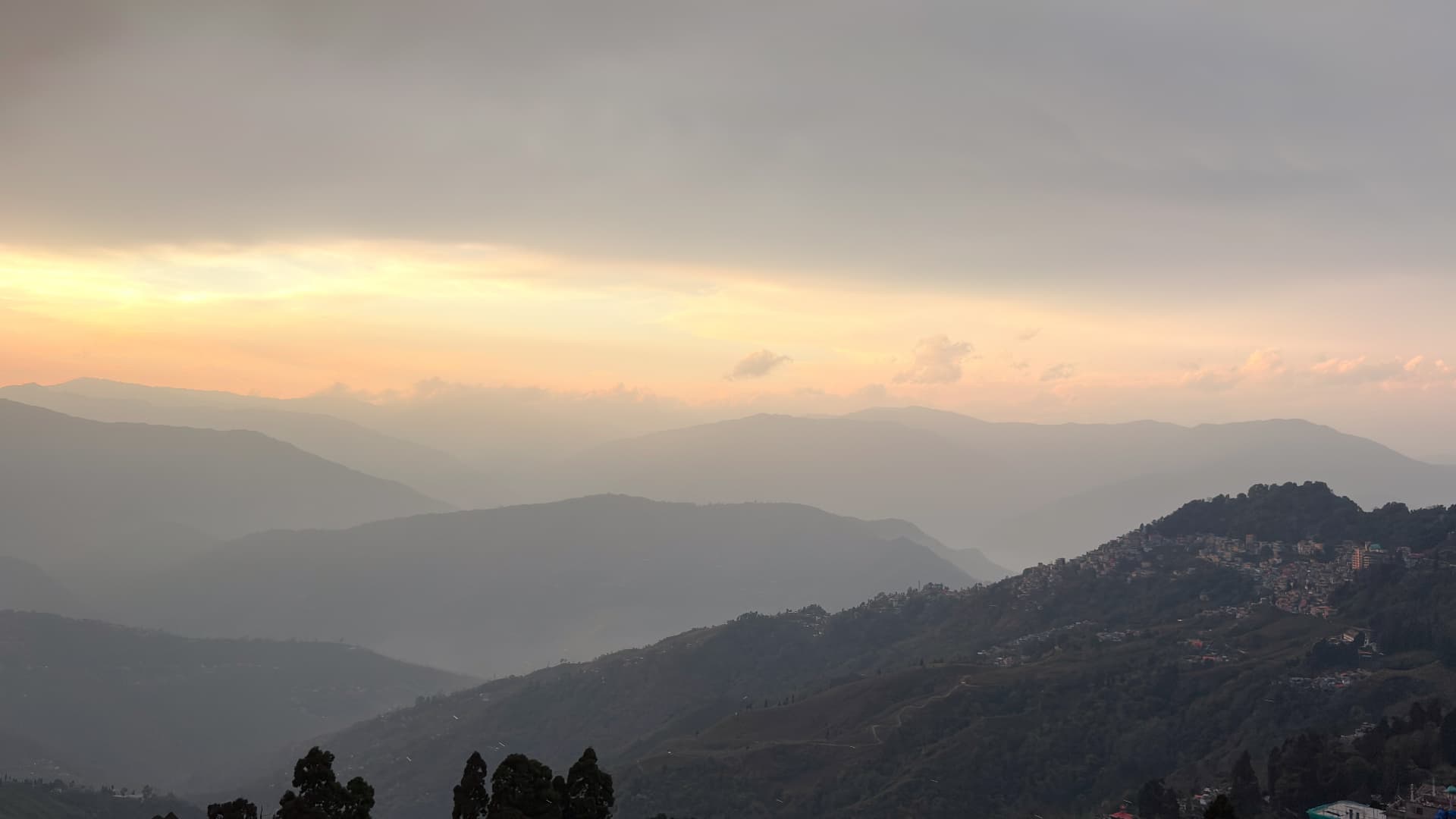A hazy sunset over layered Himalayan hills near Darjeeling, with an amber sky, silhouetted ridges receding into the distance, and a hillside town clinging to the right slope.