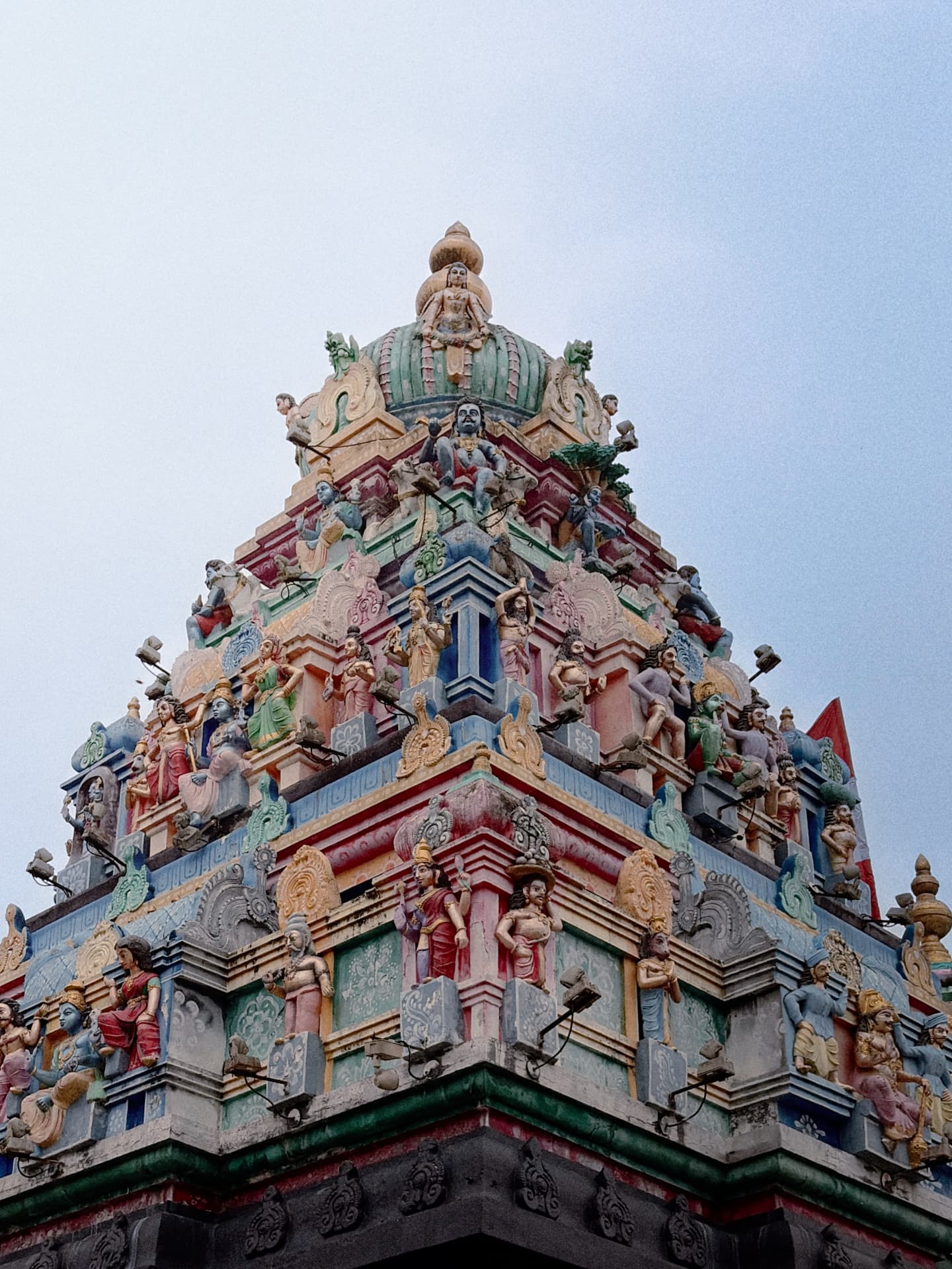 Tall colorful South Indian Hindu temple gopuram tower covered in intricately painted sculptures of gods, goddesses and mythological figures, viewed from below against a soft grey sky.